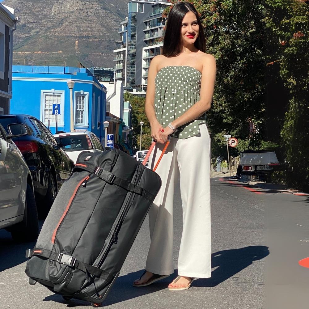 Lifestyle image of a woman holding the Dallas Roller Duffel in an outdoor café setting, emphasizing its spacious design, sturdy build, and effortlessly stylish travel vibe