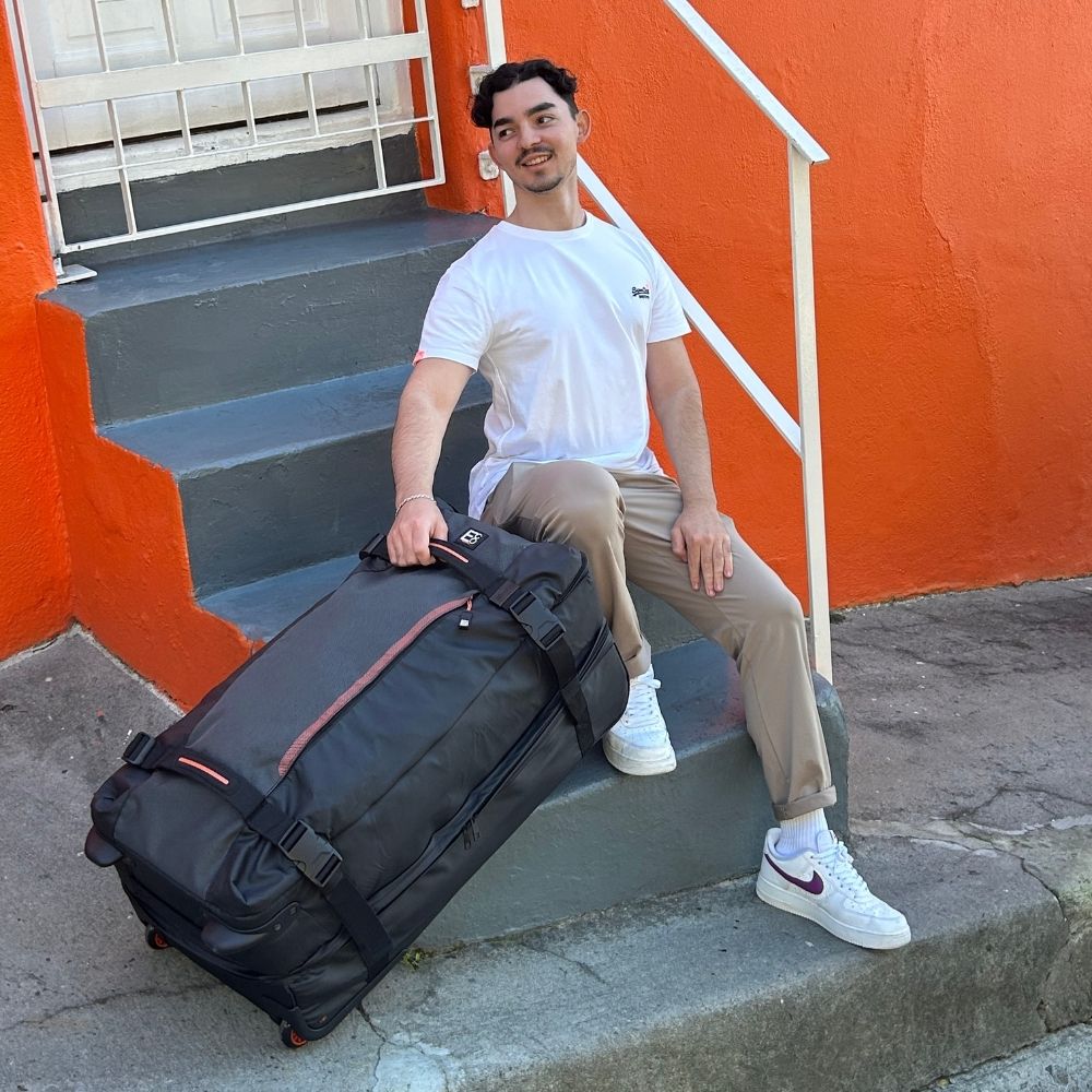 Lifestyle image of a man holding the Dallas Roller Duffel in an outdoor café setting, showcasing its durable construction, generous capacity, and modern, on-the-go functionality
